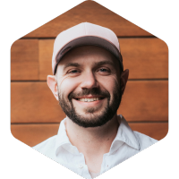 Max, beard and mustache with a hat on, smiling on a background of wood